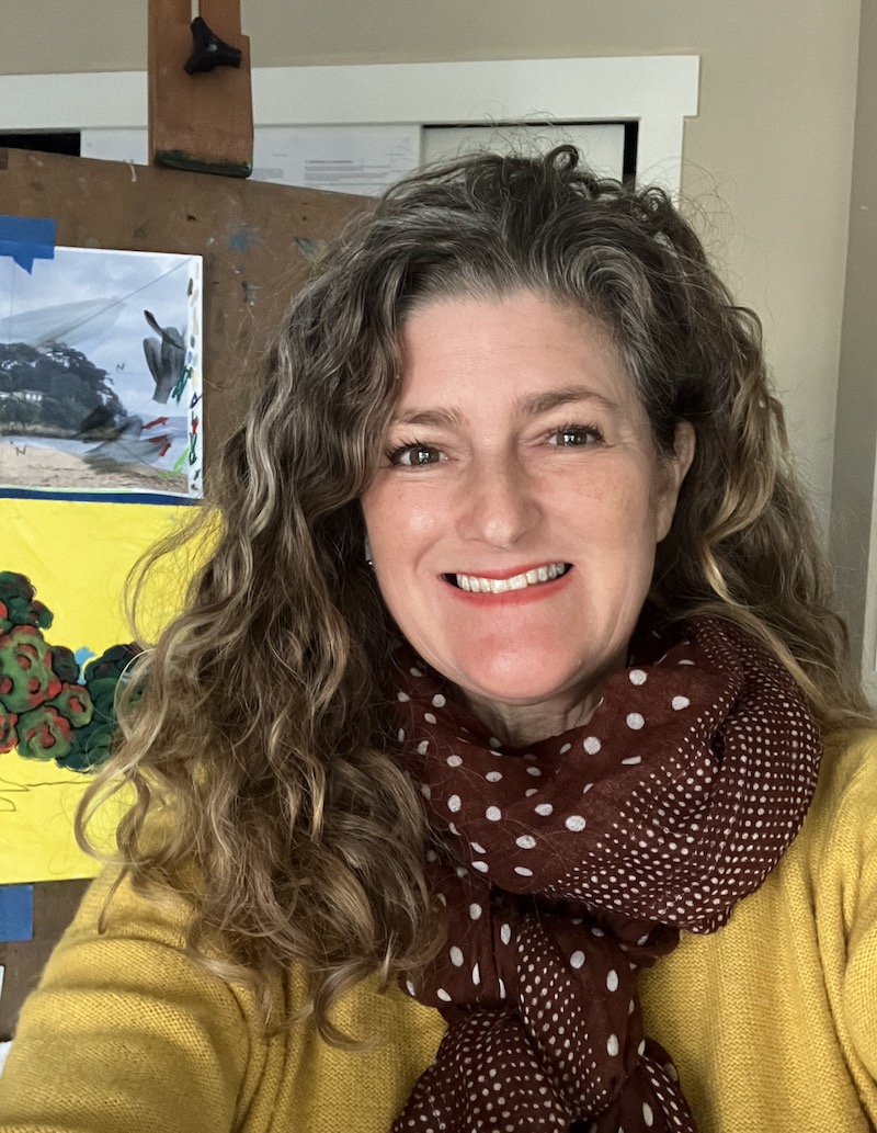 Nicola Warner artist in studio wearing yellow with a painting on the easel
